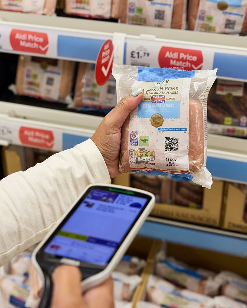 A shopper scanning the QR code on a pack of Tesco sausages with a handheld scanner in the fresh aisle